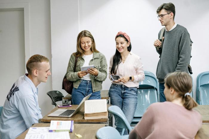 Grupa uczniów rozmawiających z prowadzącym przy biurku z laptopem i materiałami edukacyjnymi, część z nich trzyma notatniki i telefony. Ujęcie pokazuje wspólne omawianie materiału oraz naukę w małej grupie, charakterystyczną dla skutecznego przygotowania do matury w Gdańsku z Centrum Edukacyjnym Symposio.